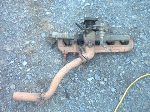 1985 Ford Bronco - A rusty exhaust/intake manifold assembly with an attached downpipe lies on gray gravel, partially coated in orange-brown rust and dirt. A y…