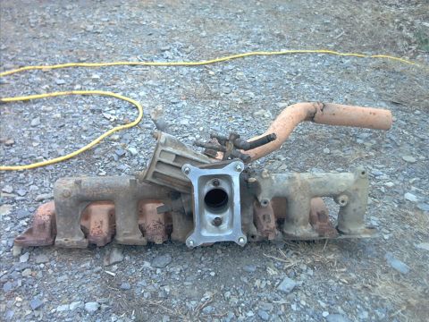 1985 Ford Bronco - A removed V8 intake manifold sitting upside down on gravel, with rusty red-brown runners and cast part numbers, a machined carburetor/throt…