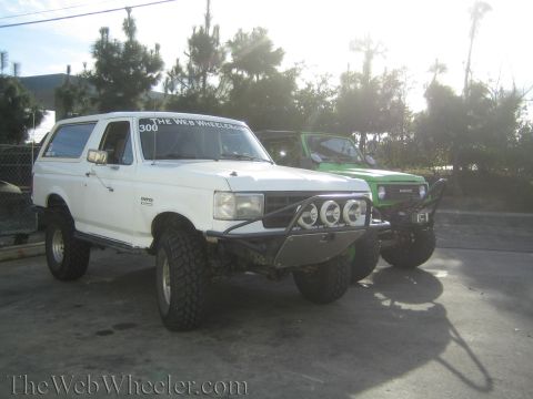 1990 Ford Bronco - white lifted SUV with front light bar parked beside a green off-road vehicle in bright backlit sunlight.