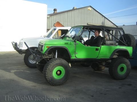 1990 Ford Bronco - bright green lifted off-road vehicle on large tires with an exposed roll cage and a seated occupant, white truck behind.