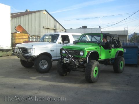 1990 Ford Bronco - green lifted Suzuki off-roader with a person at the wheel sits beside a white lifted truck outside a metal shop.