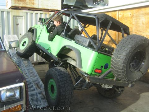1990 Ford Bronco - Rear three-quarter view of a green lifted off-road vehicle with exposed roll cage, person inside and a rear-mounted spare tire.