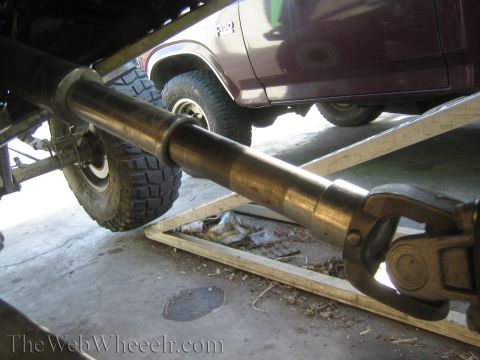1990 Ford Bronco - Underbody view of a long metal driveshaft and U-joint running beneath a maroon vehicle parked on a ramp.
