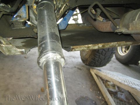 1990 Ford Bronco - Underbody close-up of a metal driveshaft passing along a vehicle chassis with blue-painted brackets and a tire visible.