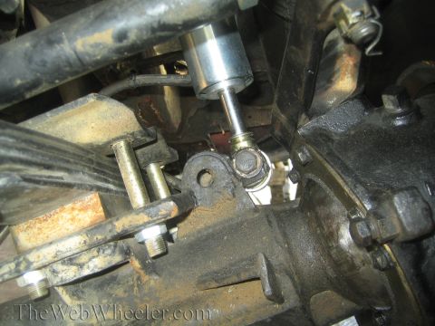 1990 Ford Bronco - Underside view of a vehicle's steering and suspension linkage showing a polished shock body, tie-rod end, and rusty hardware.