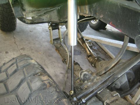 1990 Ford Bronco - Underbody view of a lifted off-road vehicle on a garage floor, showing driveshaft, steering stabilizer shock, and chunky tires.