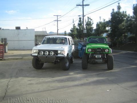1990 Ford Bronco - Two heavily lifted off-road trucks parked side by side in a paved lot, a white one with light bar and a green Suzuki.