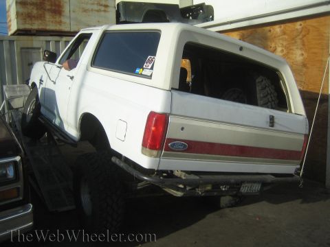 1990 Ford Bronco - White lifted Ford Bronco with a red bodyside stripe loaded on a trailer, viewed from the rear three-quarter angle.