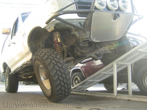 1990 Ford Bronco - Low-angle underbody view of a lifted truck on a trailer ramp, showing a coil-over shock, mud tire, and exhaust.