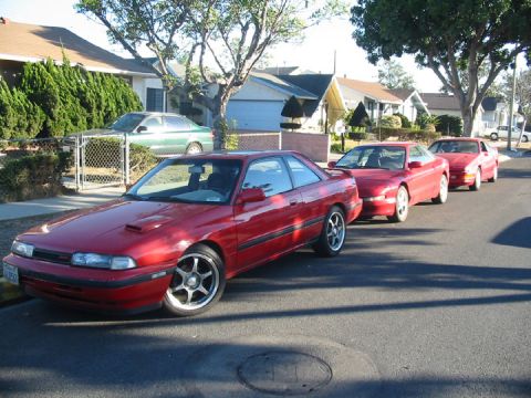 1989 Mazda MX-6 - A line of red coupes are parked on a suburban street, with houses and trees in the background.