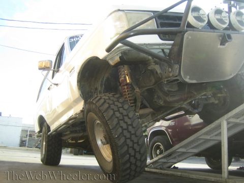 1990 Ford Bronco - Front three-quarter low view of a beige lifted truck on a trailer ramp, showing coil-over suspension and overhead light bar.