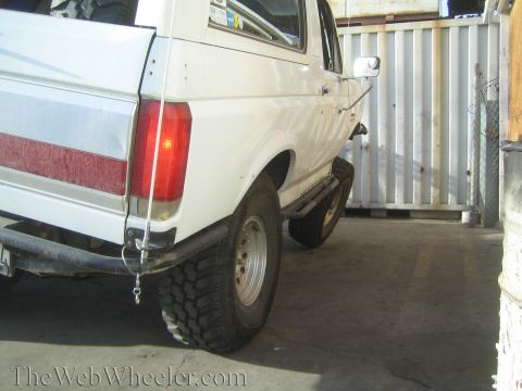 1990 Ford Bronco - Rear three-quarter view of a white lifted Ford Bronco on large tires, parked beside a corrugated metal wall.