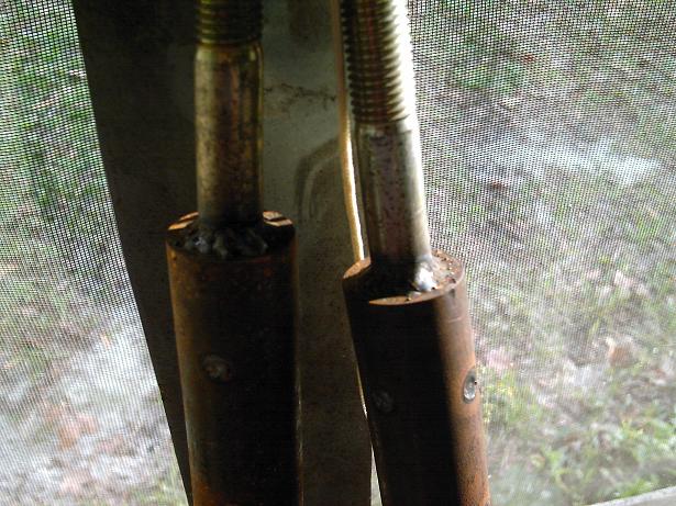 two rusty metal rods with threaded tops and bolts on the sides, seen through a screen