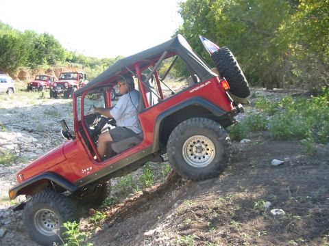 1996 Ford Bronco - A red Jeep Wrangler navigates a rocky, off-road trail with other vehicles visible in the distance.