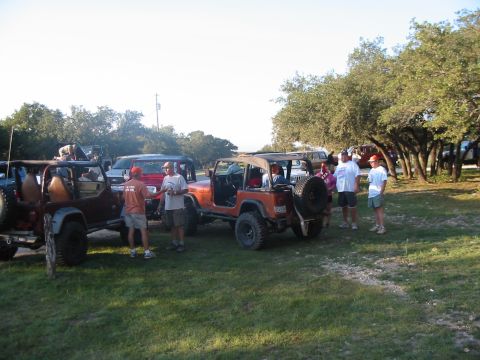 1996 Ford Bronco - Several Jeeps are gathered on a grassy area with trees in the background, and a group of people are socializing.
