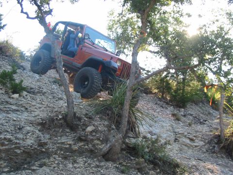 1996 Ford Bronco - An orange Jeep Wrangler navigates a steep, rocky off-road trail with trees and brush.