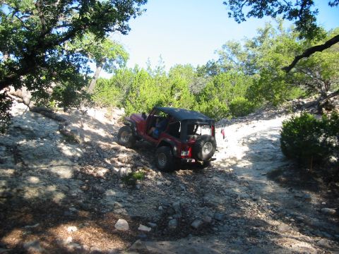 1996 Ford Bronco - A red Jeep Wrangler navigates a challenging off-road path surrounded by trees and rocky terrain.
