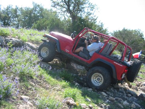 1996 Ford Bronco - A red Jeep Wrangler navigates a steep, rocky incline with sparse vegetation and wildflowers.