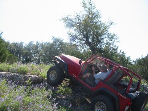 1996 Ford Bronco - A red Jeep Wrangler navigates a steep, rocky incline surrounded by greenery and wildflowers.