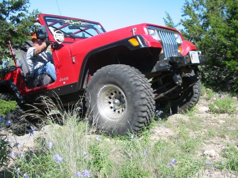 1996 Ford Bronco - A red Jeep Wrangler navigates a rocky, off-road trail with sparse vegetation and blue flowers.
