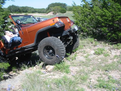 1996 Ford Bronco - An orange Jeep Wrangler navigates a rocky, off-road trail with trees on either side.