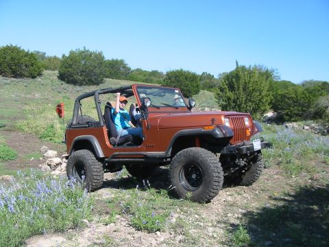 1996 Ford Bronco - A person drives an orange Jeep Wrangler on a dirt path in a grassy, hilly area with wildflowers.