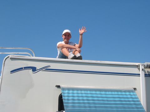 1996 Ford Bronco - A person sits on the roof of an RV, waving to the camera under a clear blue sky.