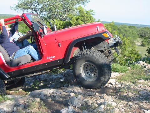 1996 Ford Bronco - A red Jeep Wrangler is shown off-roading on a rocky trail with trees in the background.
