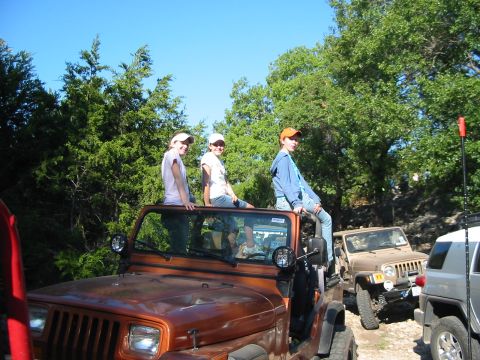 1996 Ford Bronco - Three young people sit atop an orange Jeep Wrangler, with another Jeep parked closely behind it.