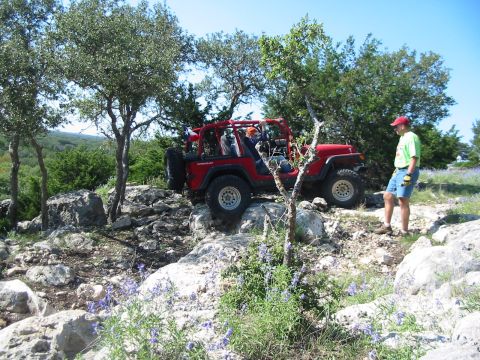 1996 Ford Bronco - A red Jeep Wrangler is shown off-roading on a rocky path, with a person standing to the side.