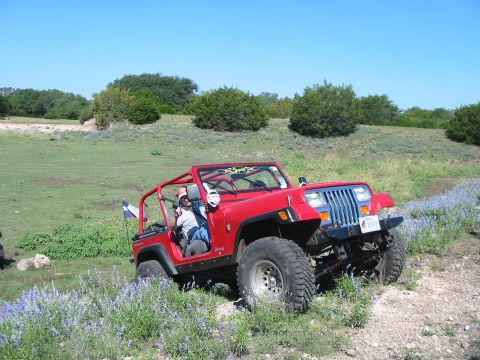 1996 Ford Bronco - A red Jeep Wrangler navigates a dirt path through a grassy, hilly landscape dotted with wildflowers under a clear blue sky.