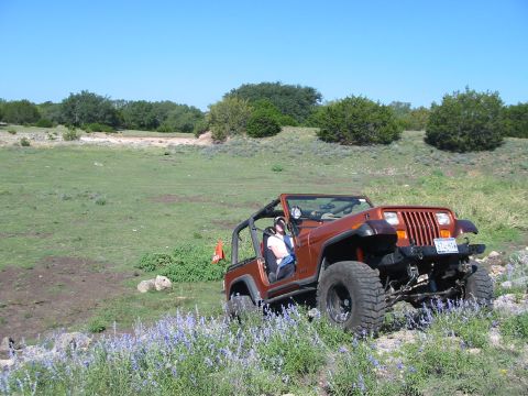 1996 Ford Bronco - A Jeep Wrangler navigates an off-road trail with a driver visible inside. The vehicle is orange and has large tires.