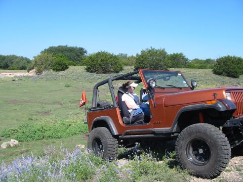 1996 Ford Bronco - A woman sits in a tan Jeep Wrangler with an orange flag on the back, parked in a grassy, outdoor area.