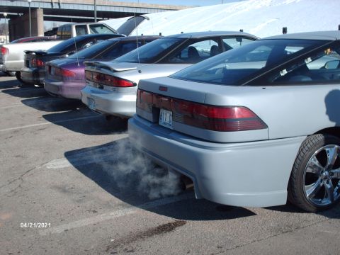 1995 Ford Probe - A line of cars, including a light gray 1995 Ford Probe emitting exhaust smoke, are parked in a lot.