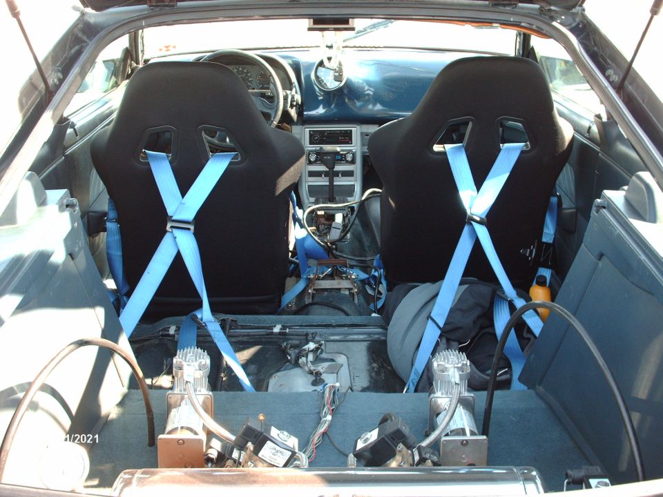 Car interior showing racing seats with blue harnesses, dashboard, and two air compressors in the rear