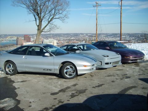 1995 Ford Probe - Three Ford Probes are parked on a snowy overlook, with a city skyline visible behind them.