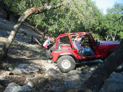 1996 Ford Bronco - A red Jeep Wrangler navigates a rocky off-road trail, with a Texas flag attached to its spare tire.