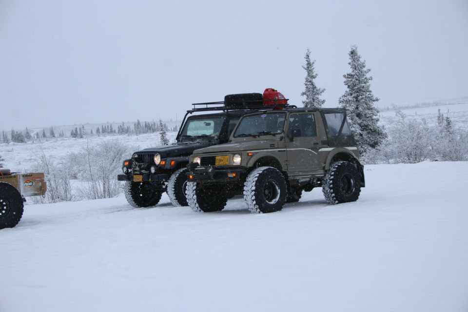 Two off-road vehicles, a black Jeep TJ and a tan Suzuki Samurai, parked in snow with trees in the background