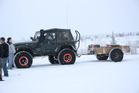 2003 Jeep TJ - A dark green Jeep Wrangler TJ is seen from the side, equipped with large off-road tires and towing a trailer through a snowy landscape.