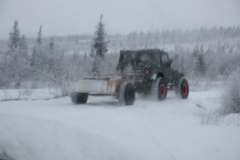 2003 Jeep TJ - A dark Jeep TJ tows a trailer through a snowy landscape, kicking up snow as it drives.