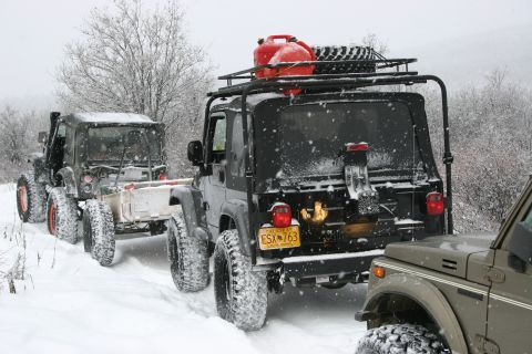 2003 Jeep TJ - Multiple Jeeps navigate a snowy trail, one towing a trailer, equipped with a roof rack carrying a gas can and spare tire.