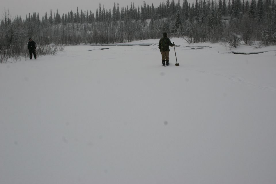 Two people stand in a vast snowy field with a treeline behind them