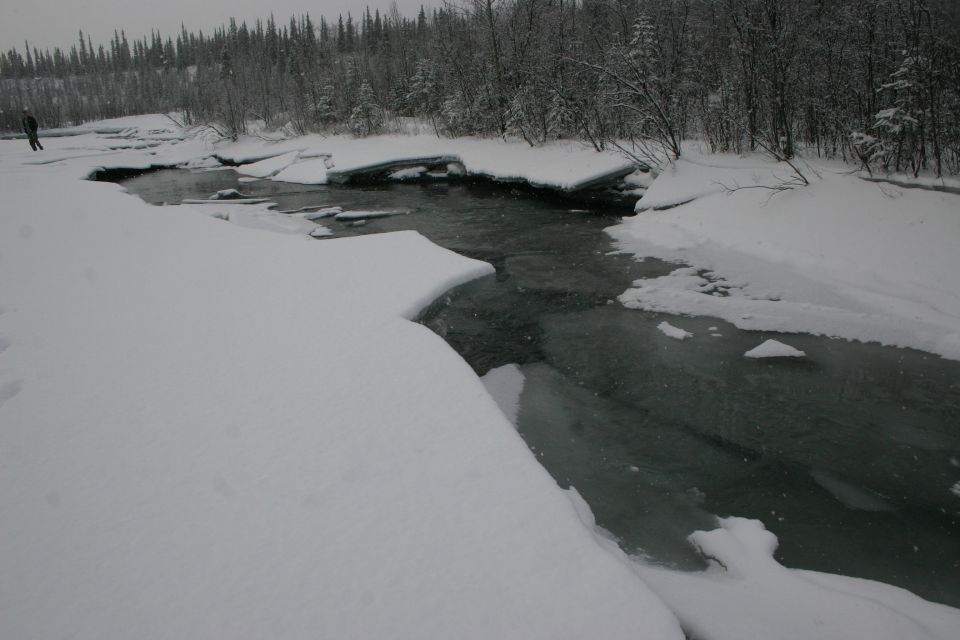 Person stands on snow-covered bank beside a dark, partially frozen river with ice floes and snow-covered trees in the background