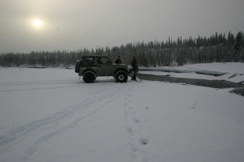 2003 Jeep TJ - An olive green SUV is parked on a snow-covered riverbank, with two individuals standing beside it as the sun sets.