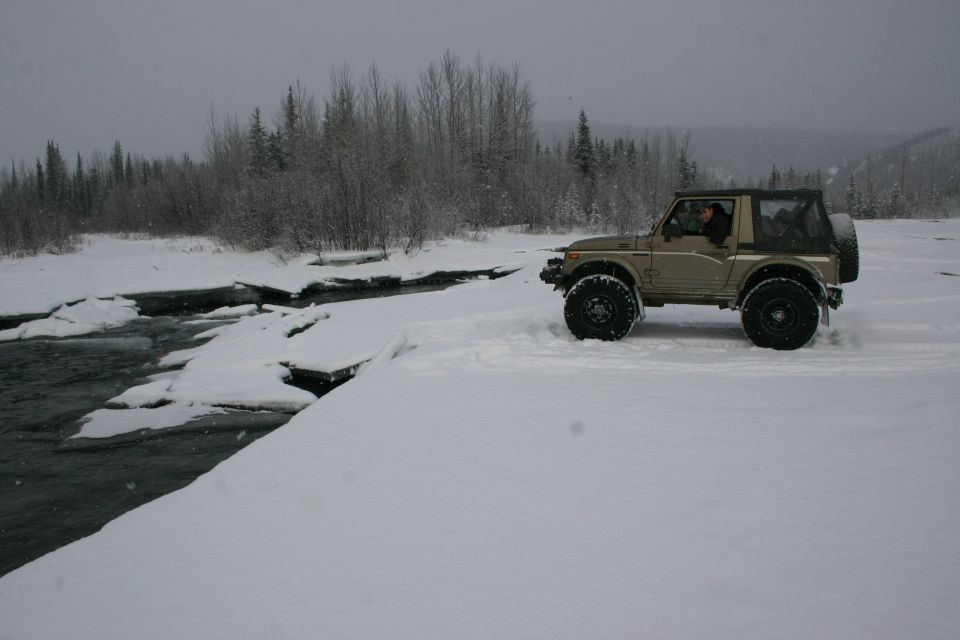 Tan Suzuki Samurai driving on a snowy path next to a partially frozen river