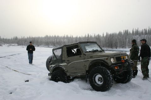 2003 Jeep TJ - A Suzuki Samurai is stuck in deep snow near a treeline with several people around it.