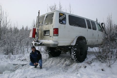 2003 Jeep TJ - A white van modified with large tires sits in a snowy landscape, with a person crouching nearby.