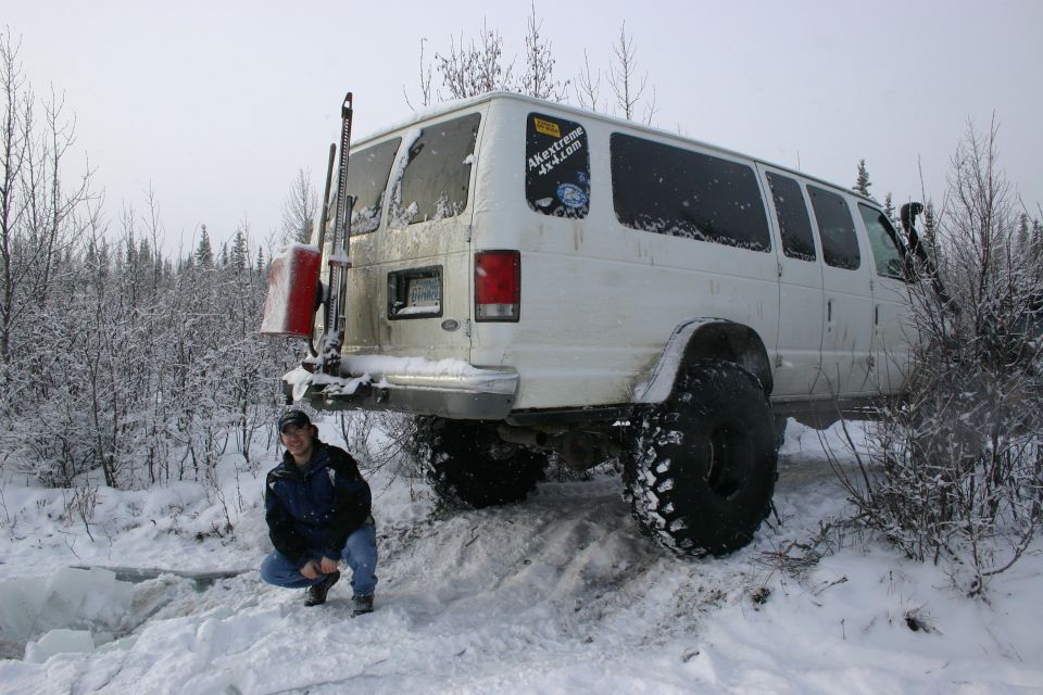 White van with oversized tires and a shovel mounted on the rear bumper, parked in a snowy, wooded area