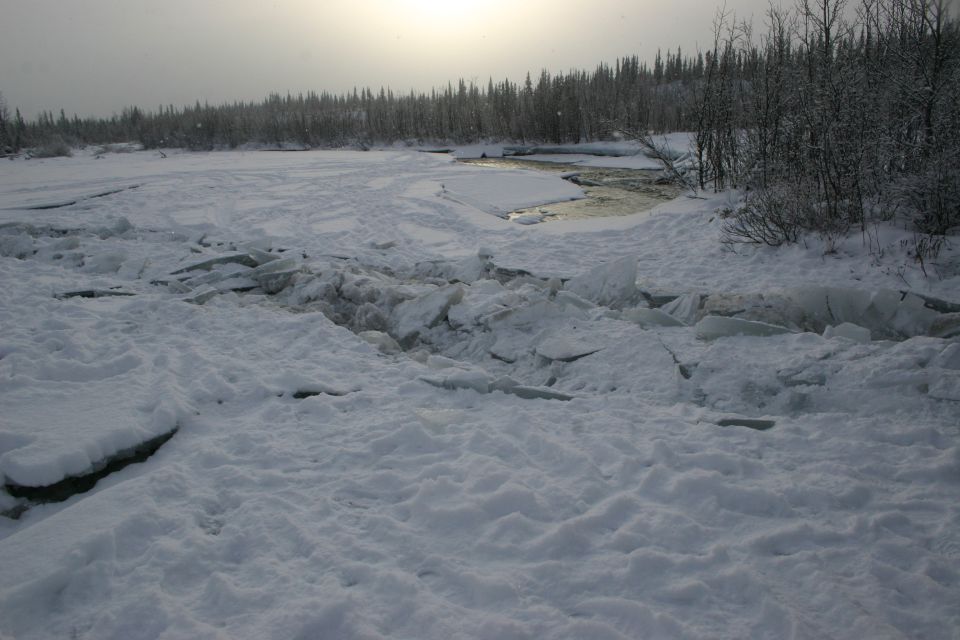 Broken ice chunks piled up on a partially frozen river with trees in the background under a hazy sky
