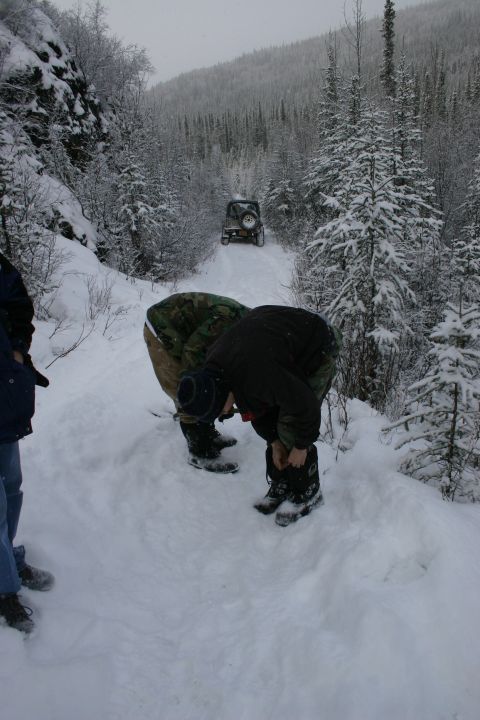2003 Jeep TJ - Two people are adjusting their boots on a snowy trail while a Jeep Wrangler drives ahead in the distance.